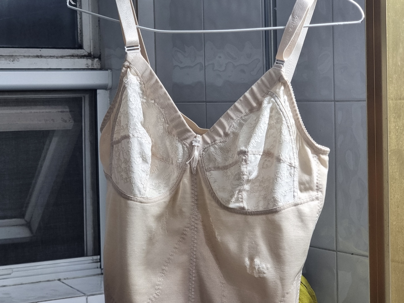 An all-in-one shapewear garment hanging on a hanger in a bathroom, looking clean but aged.